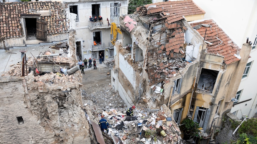 A wide aerial shot of two stone buildings and a large gap in between, amid piles of rubble and collapsed roofs and tiles.