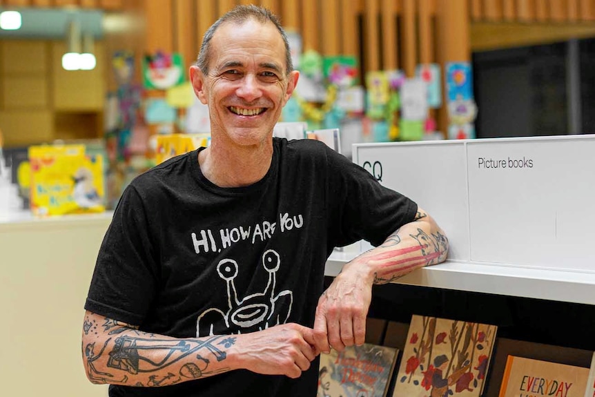 Andy smiles at the camera, leaning on a book shelf.