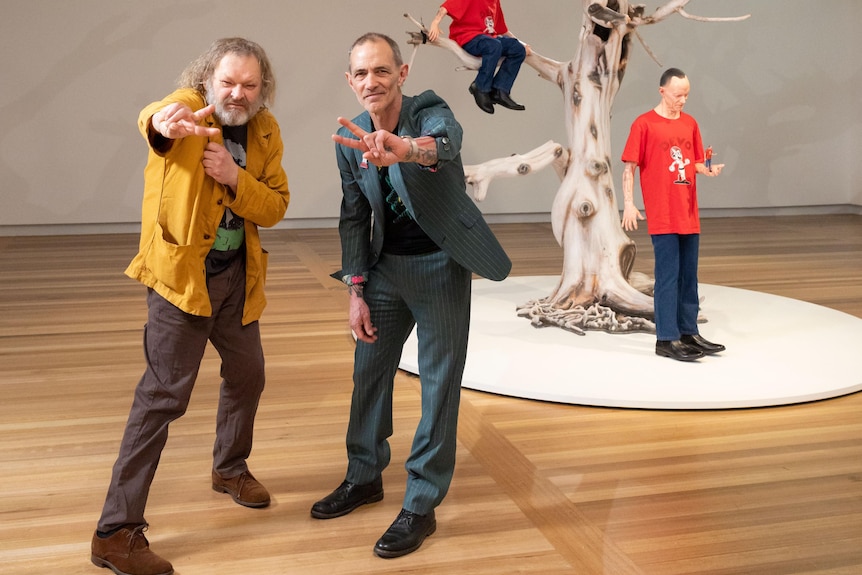 Two men stand flashing peace signs in front of a surreal sculpture of a man in a tree.