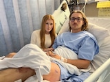 A man lies in a hospital bed with a woman beside him
