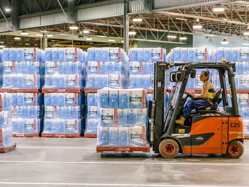 A woman drives a forklift inside a warehouse filled with toilet paper