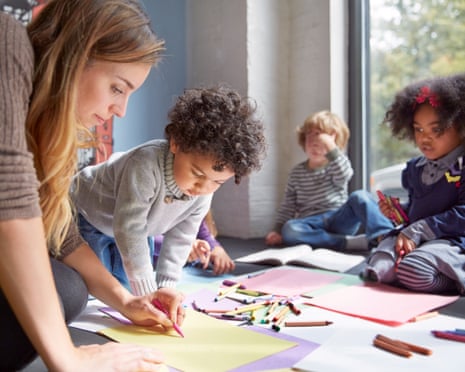 Stock image of teacher and young children sitting on a floor drawing with crayons on colored paper.